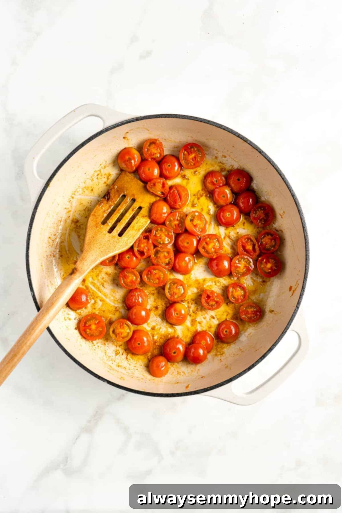 Overhead view of cooked cherry tomatoes and wooden spatula in double handled white cooking pot