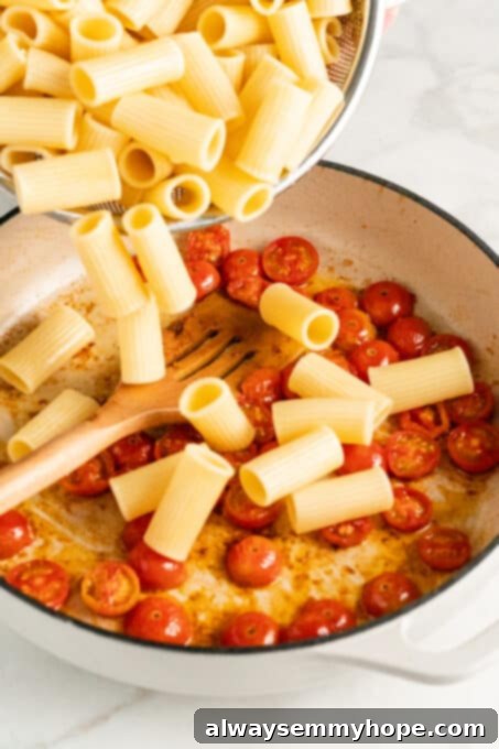 Cooked rigatone pasta being dumped from metal colander into pan with halved cherry tomatoes and wooden spatula