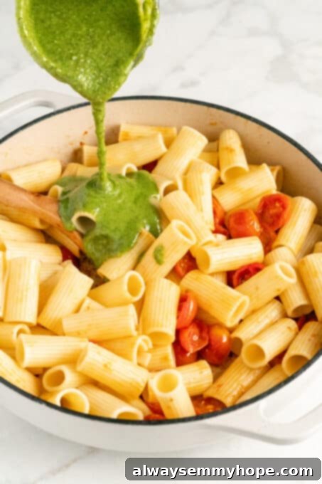 Pesto sauce being poured onto cooked rigatone pasta and cherry tomatoes inside of a double handled white cooking pot