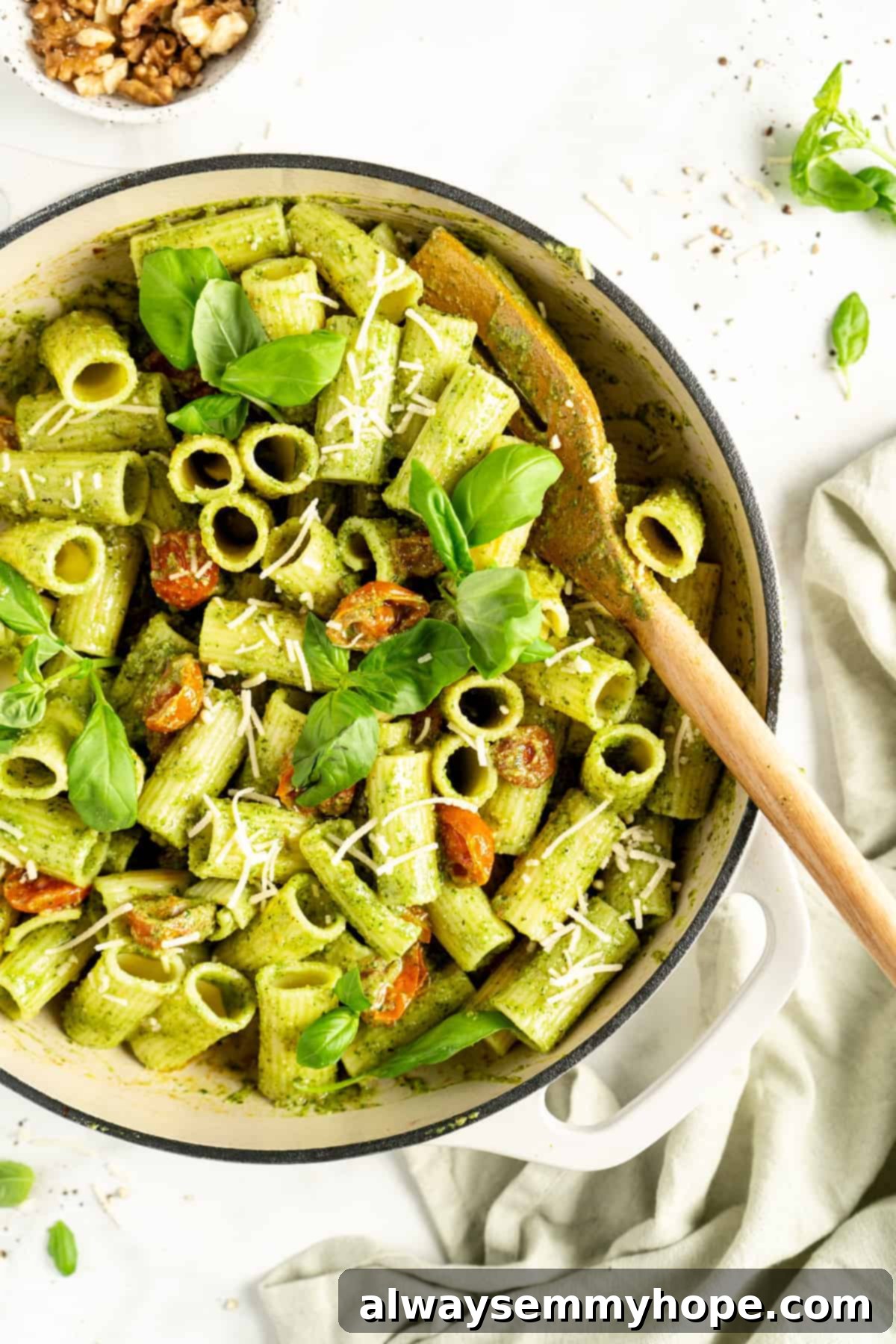 Overhead view of pesto-coated rigatone pasta, cherry tomatoes, fresh basil, and vegan parmesan cheese in round white cooking pot