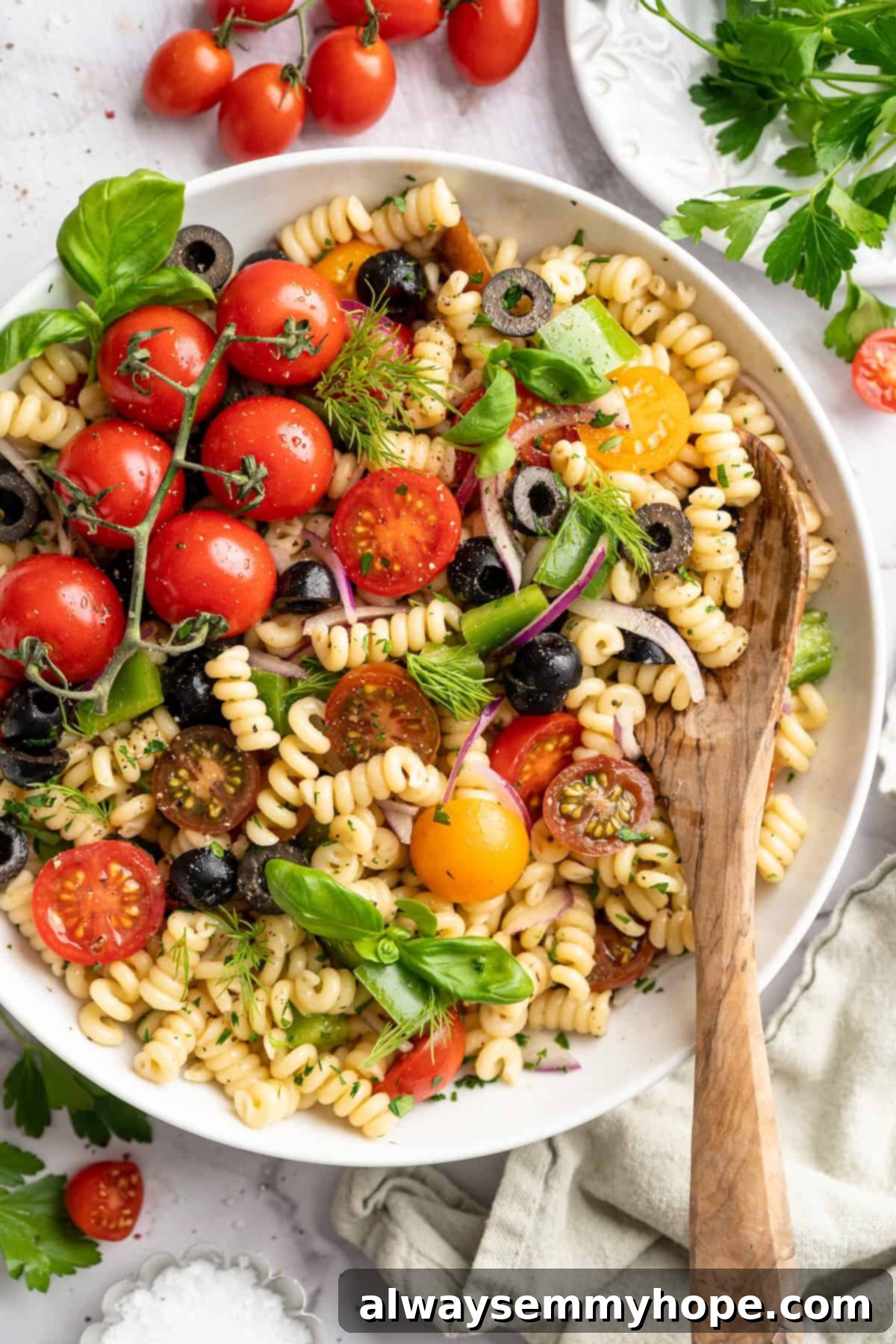 Serve this vibrant vegan pasta salad. Overhead view of a vibrant vegan pasta salad in a white serving bowl, with a wooden spoon, showcasing its rich colors and fresh ingredients.