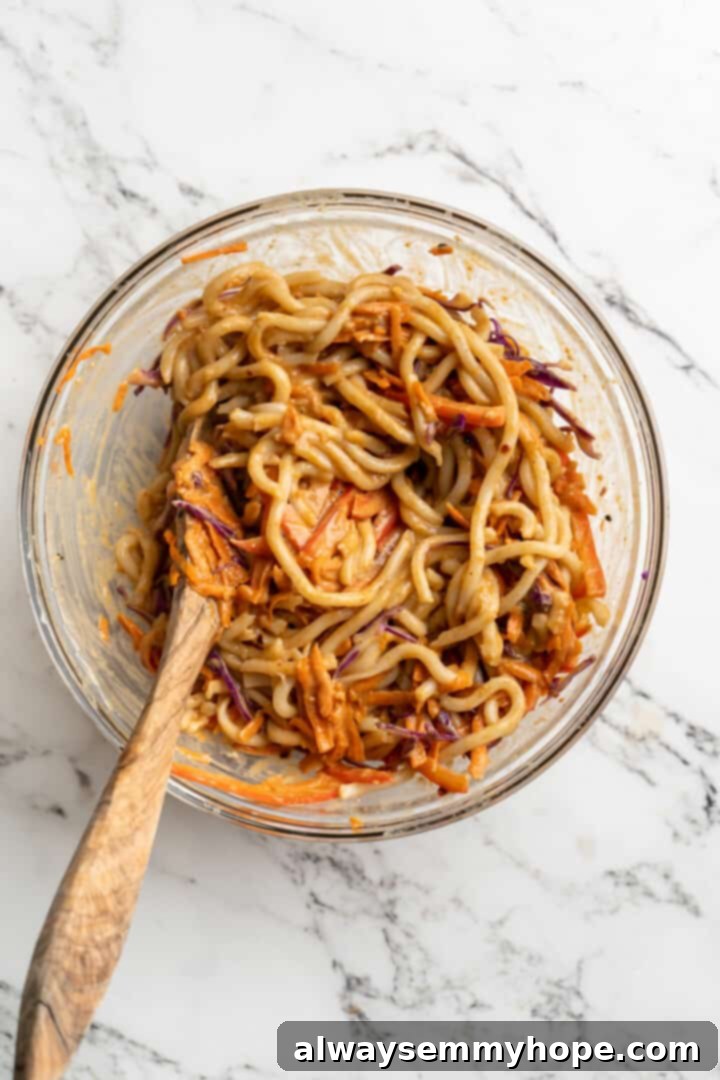 Overhead view of Thai peanut noodles in glass bowl with wooden spoon stirring