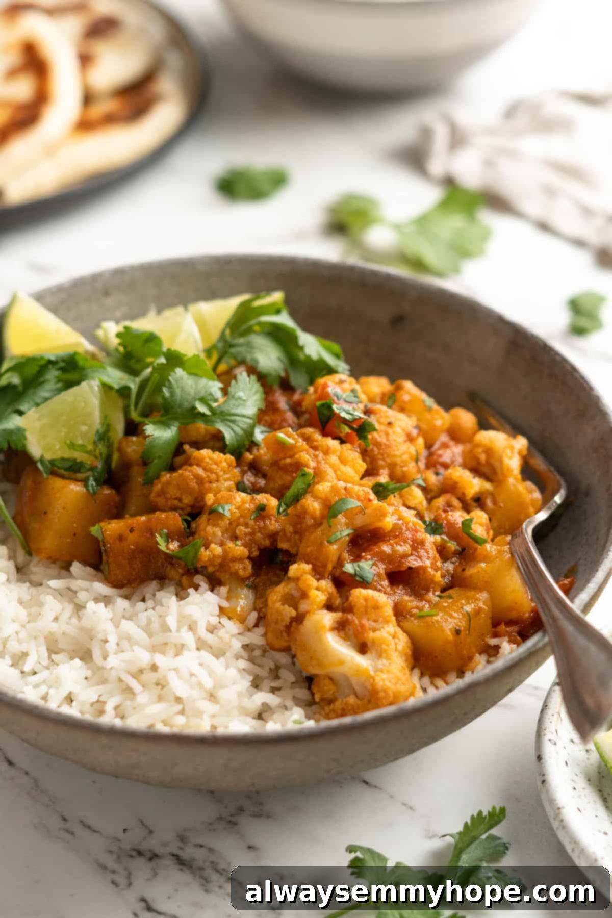 A bowl of rich Aloo Gobi, an Indian cauliflower and potato curry, artfully garnished with fresh cilantro and a lime wedge, served with a side of steamed rice.