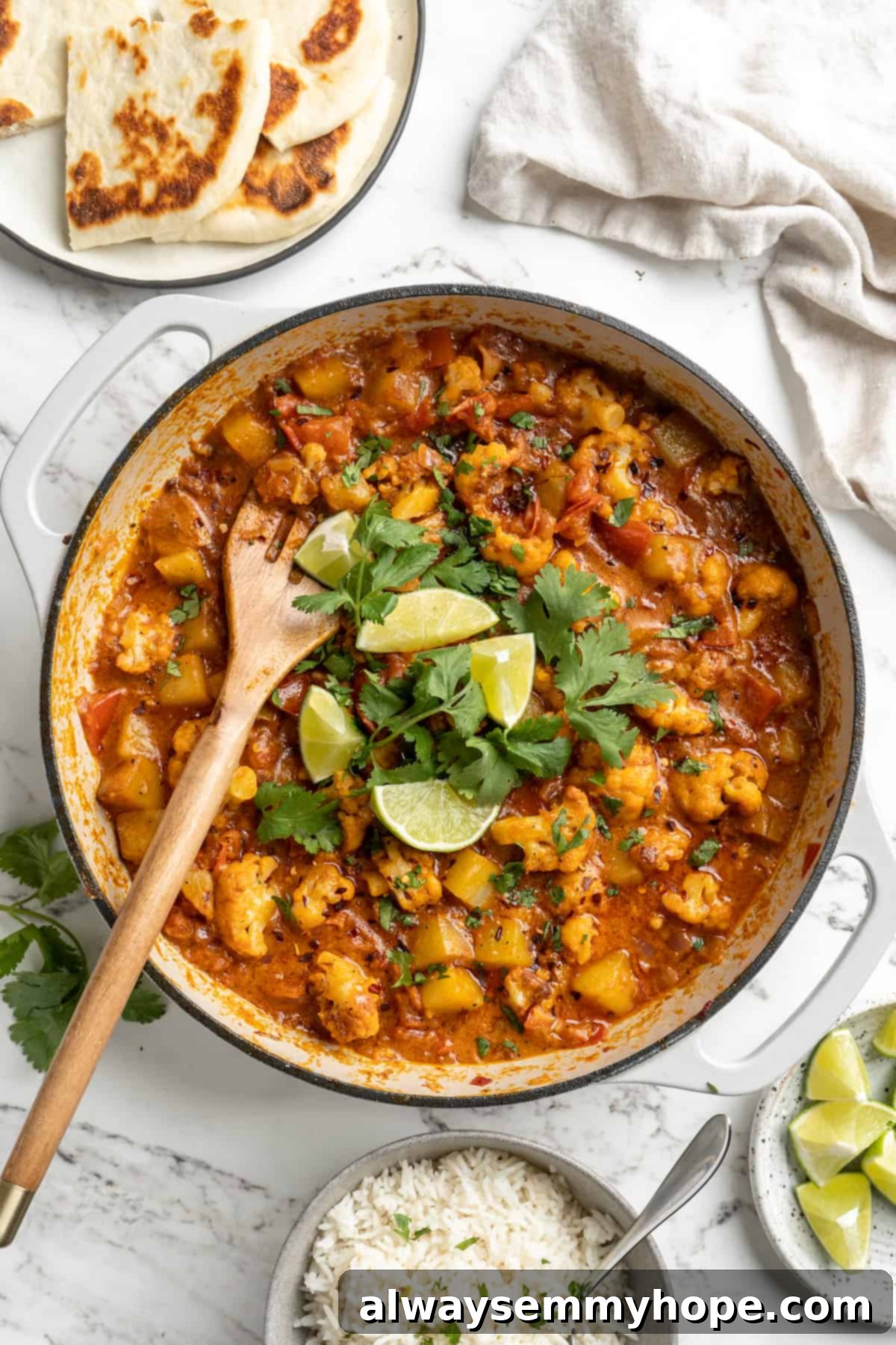 A pot of freshly cooked Aloo Gobi, a rich cauliflower and potato curry, with a wooden spoon, ready to be served. Garnished with fresh cilantro and lime wedges.