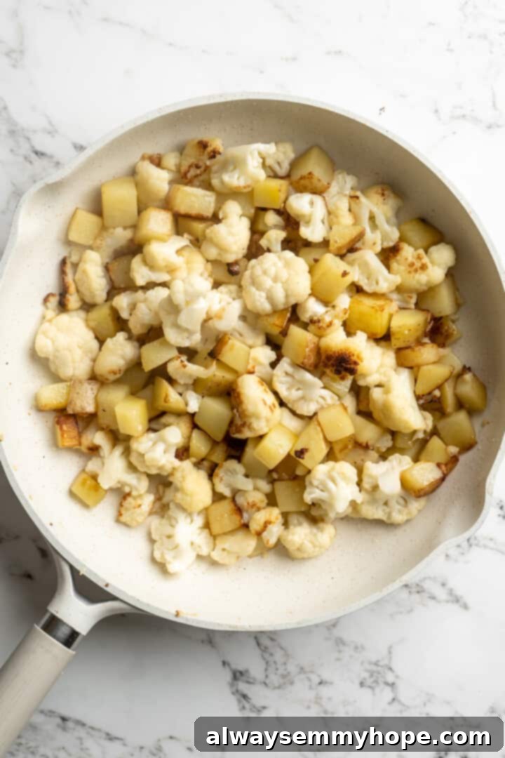Cauliflower florets and cubed Irish potatoes cooking in a large skillet with coconut oil, beginning to crisp and brown.