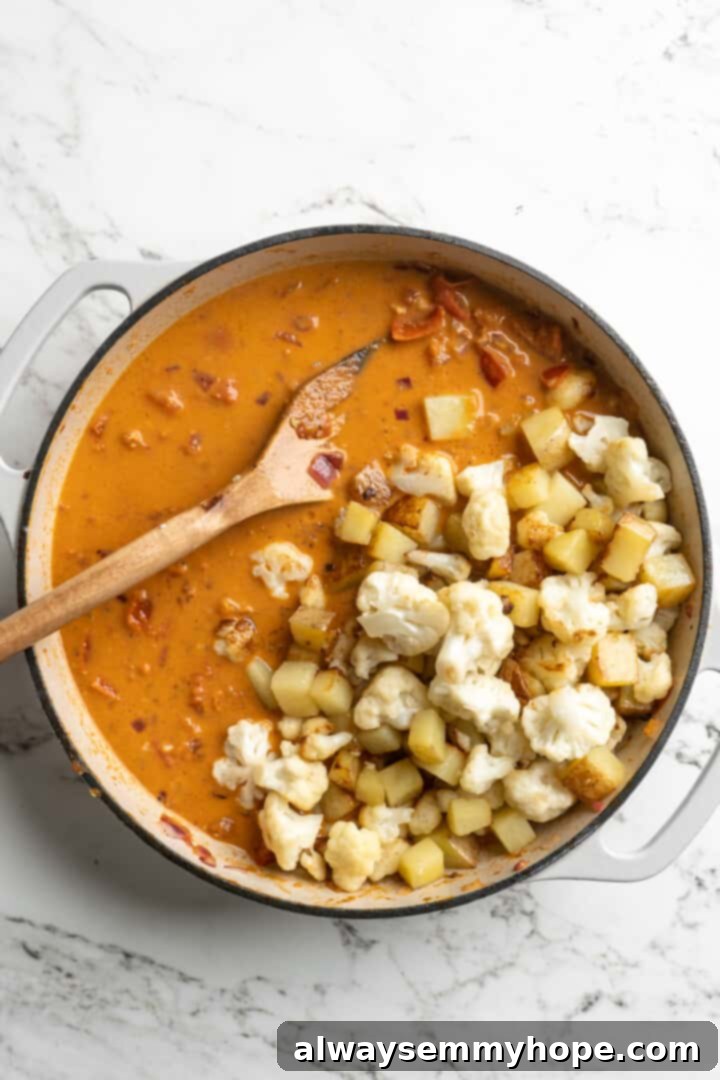 Seared cauliflower florets and cubed potatoes being added to the rich, aromatic curry sauce in a pot, ready to simmer and absorb flavors.