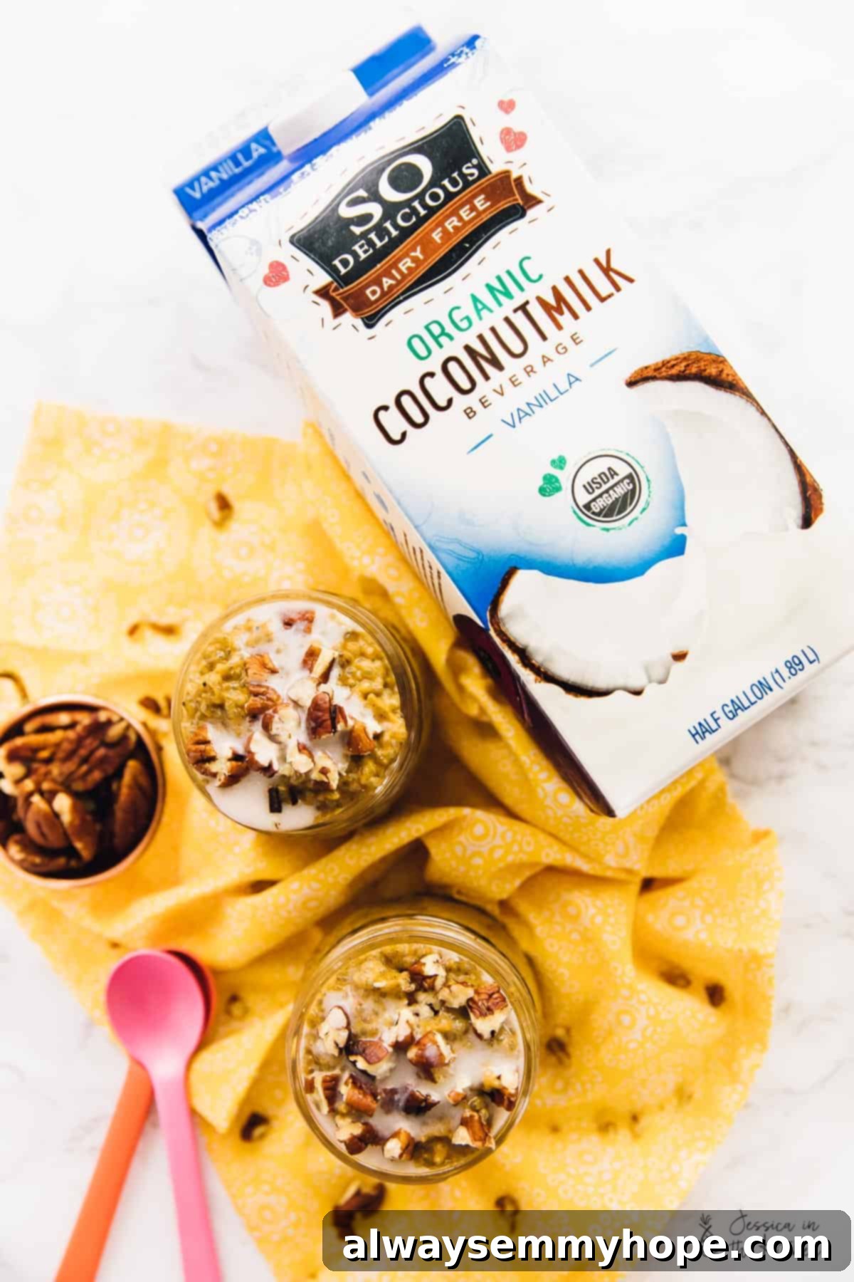 Easy vegan pumpkin chia pudding is rich, creamy, and full of fall flavors like hearty pumpkin, warm spices, and buttery pecans. Overhead view of two mason jars with chia pudding topped with pecans, next to a bowl of pecans, two spoons, and a carton of coconut milk beverage.