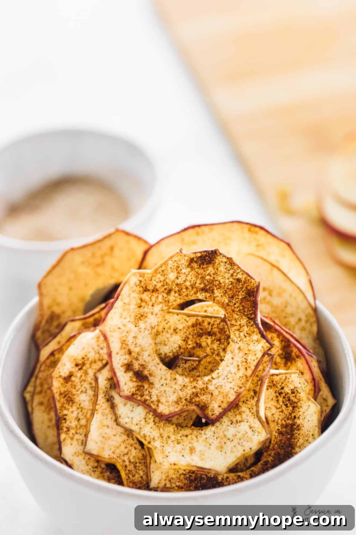 Easy homemade apple chips are crispy, chewy, richly sweet, and covered in spicy cinnamon, making them the perfect fall snack for any occasion. Close up of a bowl of golden-brown apple chips covered in cinnamon sugar, showcasing their crispy texture.