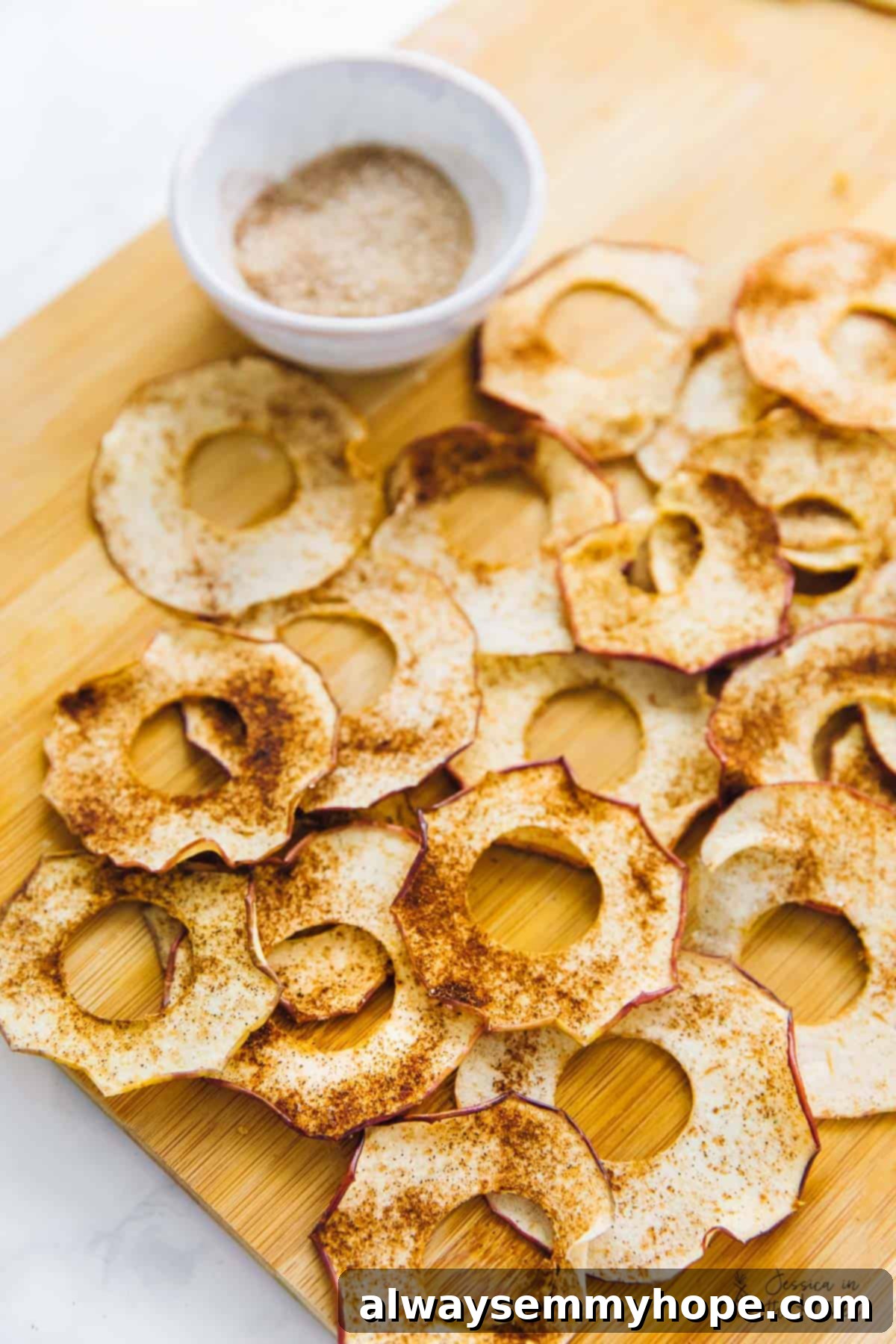 Easy homemade apple chips are crispy, chewy, rich, sweet, and covered in spicy cinnamon, making them the perfect fall snack. Overhead view of a generous pile of cinnamon apple chips on a wooden cutting board, with a small bowl of cinnamon sugar beside them.