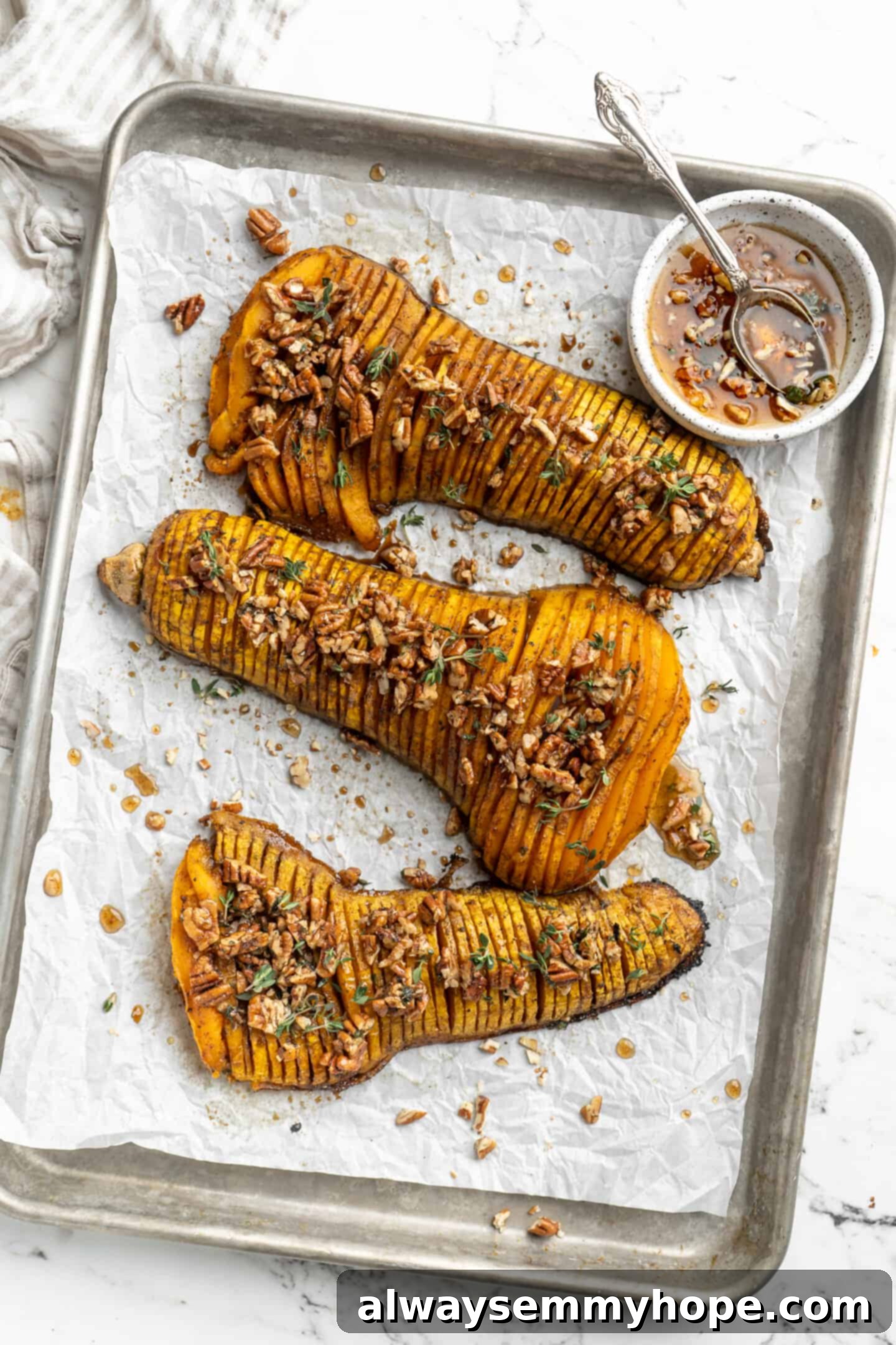 Hasselback butternut squash is a show-stopper, but it’s not all looks! Maple, tender squash, and caramelised pecans make it irresistible! Overhead view of hasselback butternut squash baking on a parchment-lined baking sheet, with a small bowl of maple pecan glaze on the side