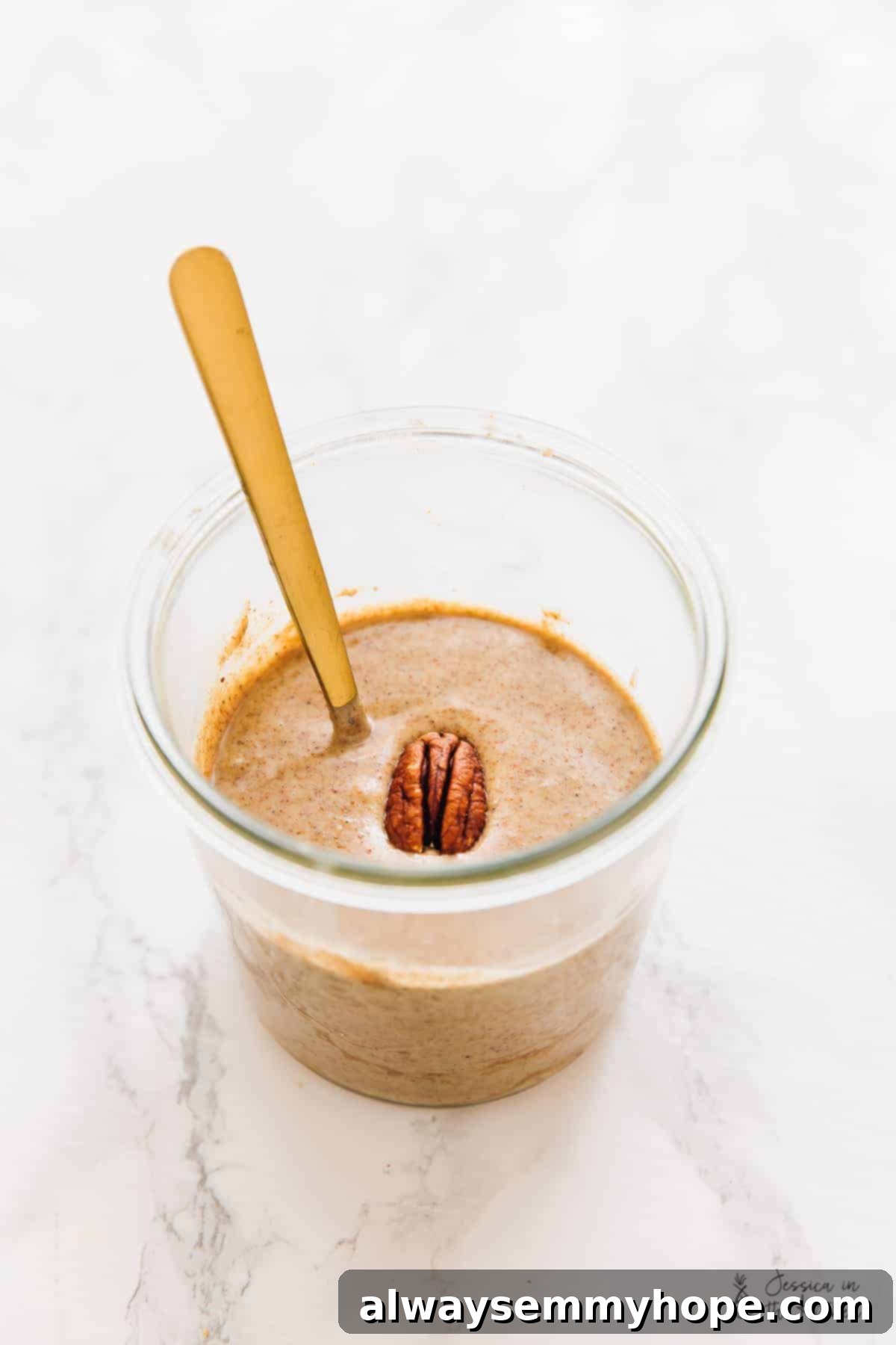 Golden Maple Pecan Sugar Cookies 4 A clear glass jar filled with rich, homemade pecan butter, with a small golden spoon resting elegantly beside it on a light background.