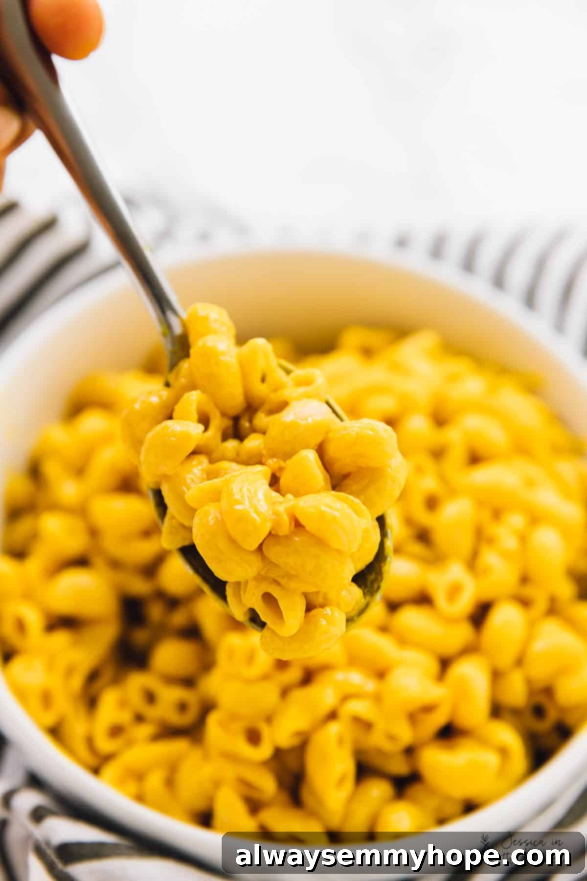 A hand holding a spoonful of mac and cheese, with a bowl of mac and cheese in the background.