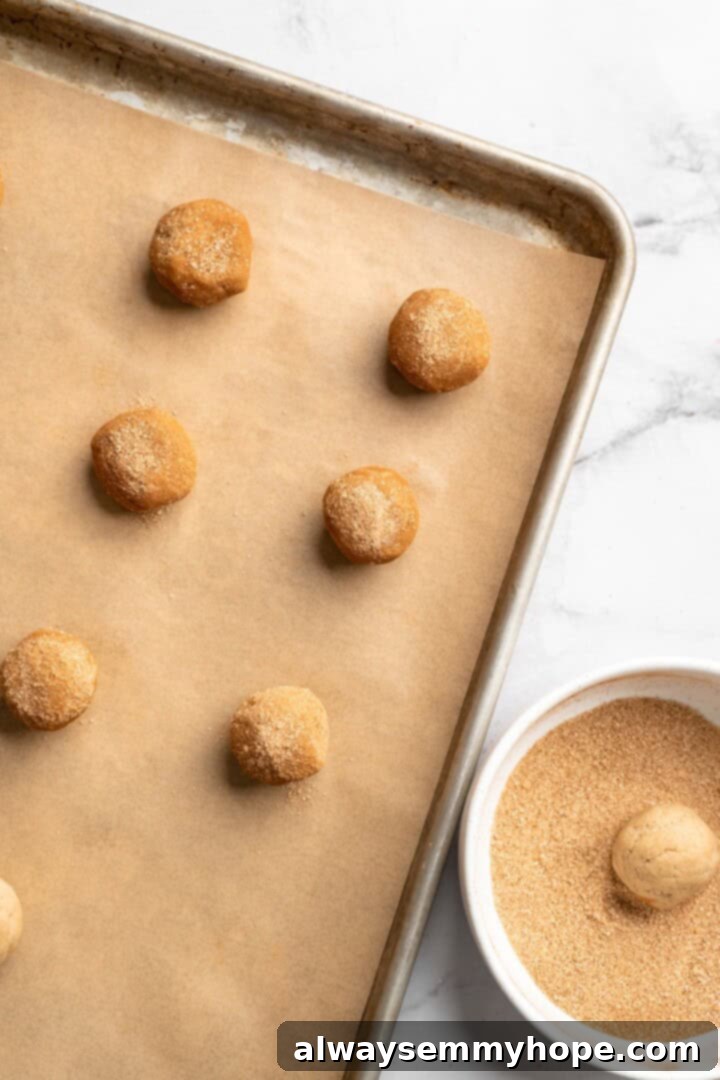 Overhead view of snickerdoodle dough balls being rolled in cinnamon sugar, with some already placed on a baking sheet.