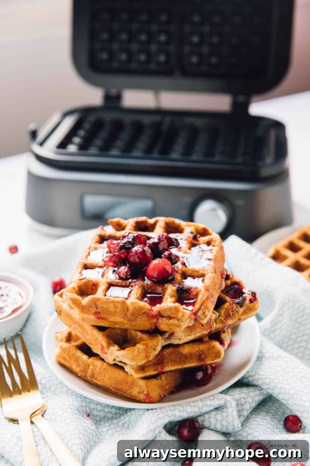 A tempting stack of golden-brown orange cranberry waffles in the foreground, with the high-tech waffle iron visible in the soft background, inviting a delicious breakfast.