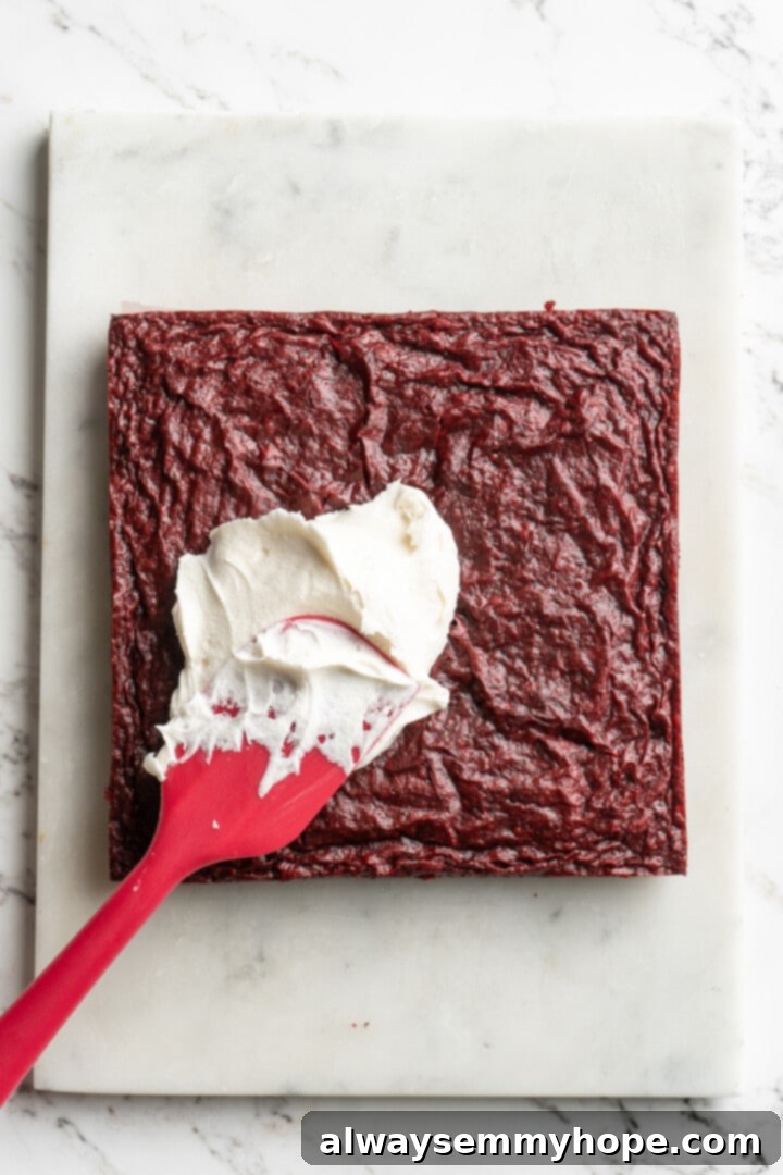 Moist, rich red velvet brownies are perfect for a special occasion or anytime you’re craving a decadent dessert. With cream cheese frosting! A spatula smoothly spreading generous layers of creamy white vegan cream cheese frosting onto cooled, vibrant red velvet brownies in a square baking pan.