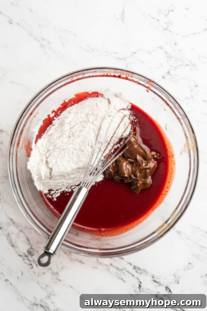 Moist, rich red velvet brownies are perfect for a special occasion or anytime you’re craving a decadent dessert. With cream cheese frosting! Overhead view of melted chocolate and the dry flour mixture carefully added to the vibrant red liquid ingredients for red velvet brownies, ready to be combined.