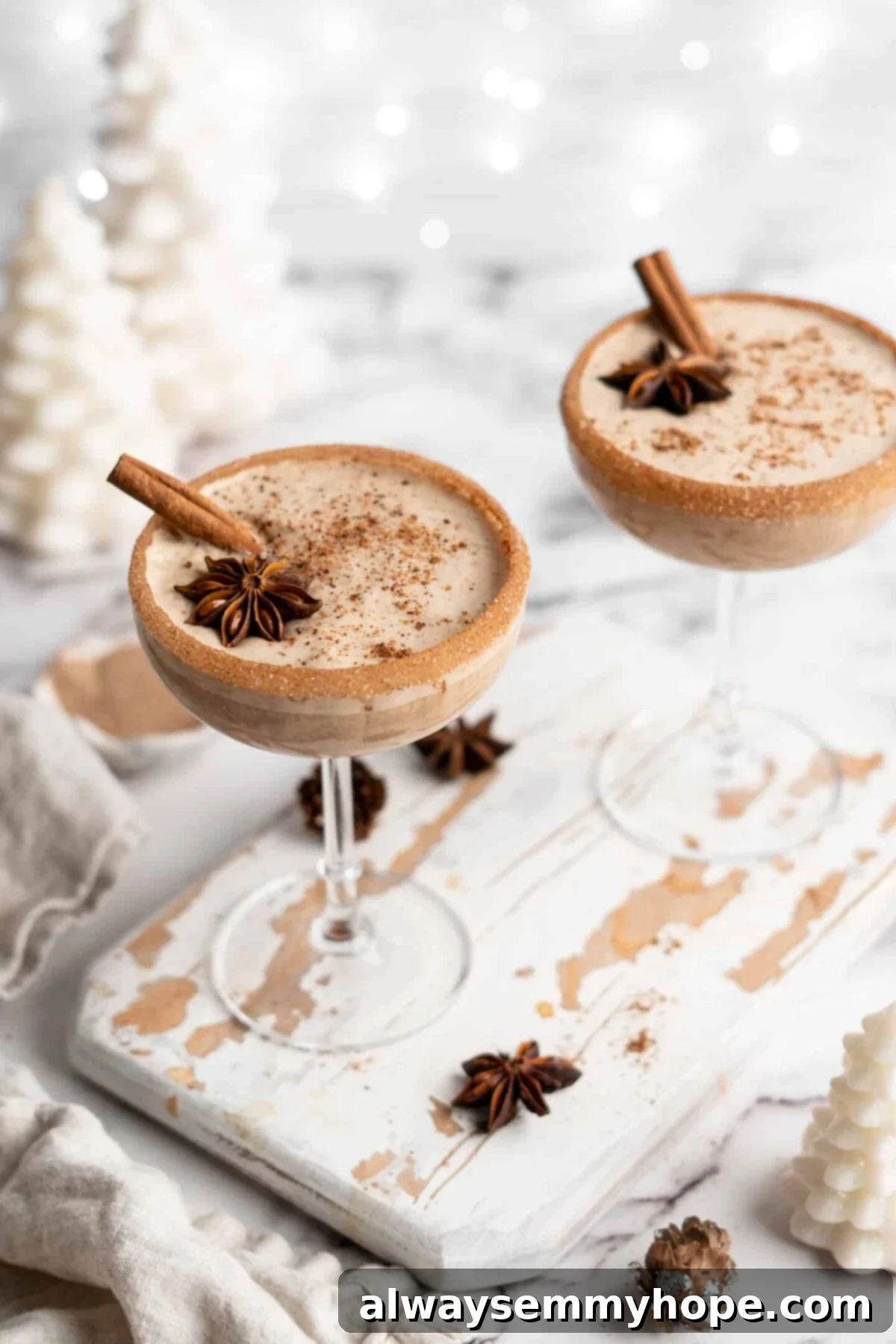 This vegan eggnog recipe is just as rich, creamy, and delicious as traditional eggnog. It's a perfect Christmas treat hot or cold! Overhead view of two cocktail glasses with vegan eggnog, topped with ground cinnamon, whole cinnamon sticks, and whole star anise, with cinnamon sugar rims, surrounded by star anise and white Christmas decorations