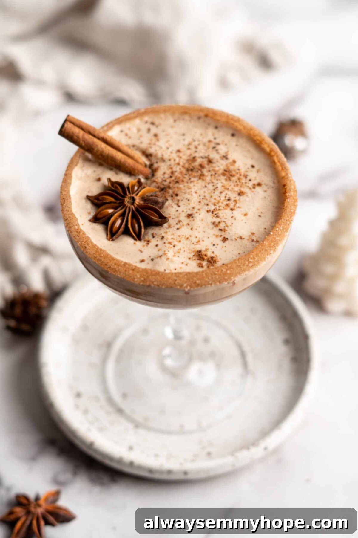 This vegan eggnog recipe is just as rich, creamy, and delicious as traditional eggnog. It's a perfect Christmas treat hot or cold! A cocktail glass on a plate filled with vegan eggnog, garnished with a cinnamon stick, a star anise pod, and ground cinnamon, with a cinnamon sugar rim