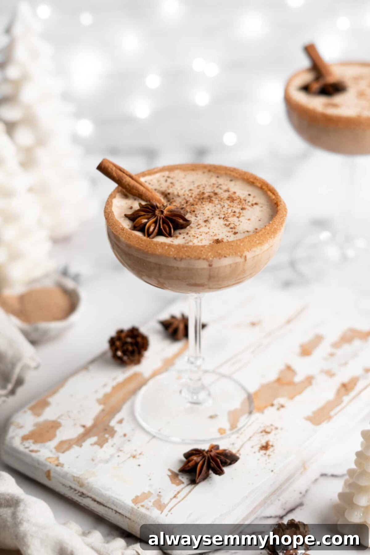 This vegan eggnog recipe is just as rich, creamy, and delicious as traditional eggnog. It's a perfect Christmas treat hot or cold! A cocktail glass filled with vegan eggnog, garnished with a cinnamon stick, ground cinnamon, and a star anise pod, with a cinnamon sugar rim, with Christmas lights and another glass of eggnog in the back