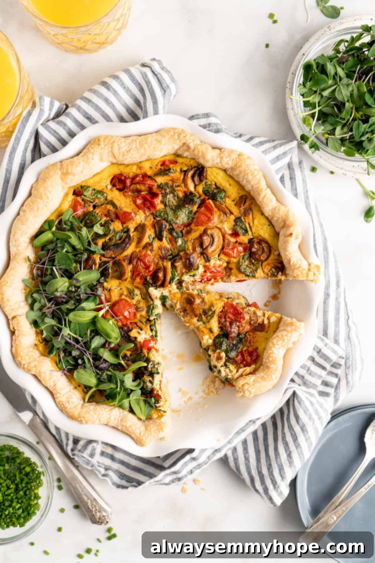 An overhead shot of a freshly baked vegan quiche in a pie dish, with bowls of vibrant green spinach and chopped chives in the background, ready for garnishing and serving.