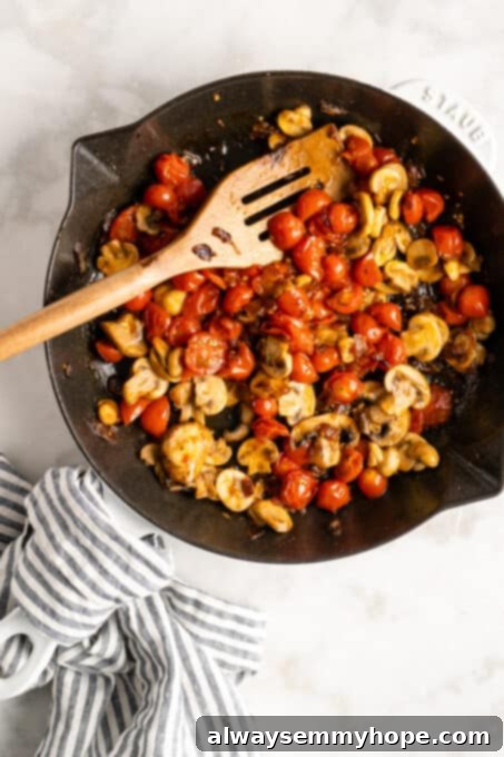 An overhead view of colorful cherry tomatoes and sliced mushrooms sizzling in a cast iron skillet alongside aromatic garlic, beginning to soften and release their flavors.