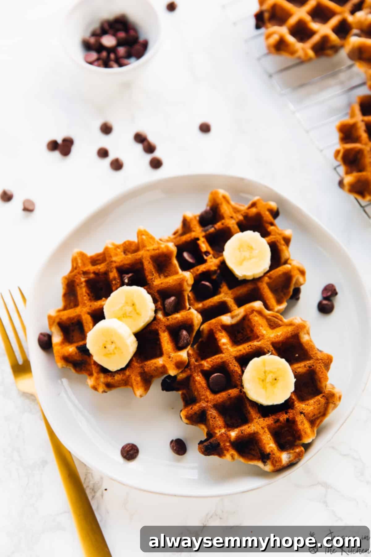 An overhead view of three perfectly cooked banana bread waffles on a white plate, accompanied by a stylish gold fork, inviting you to savor every bite.