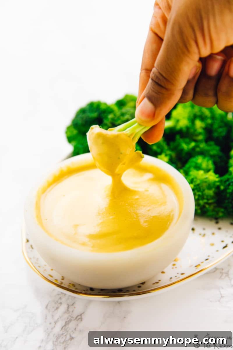 A hand gently dips a vibrant green piece of broccoli into a small bowl of creamy vegan cheese sauce, with a larger plate of broccoli florets in the background, highlighting a healthy and delicious snack option.
