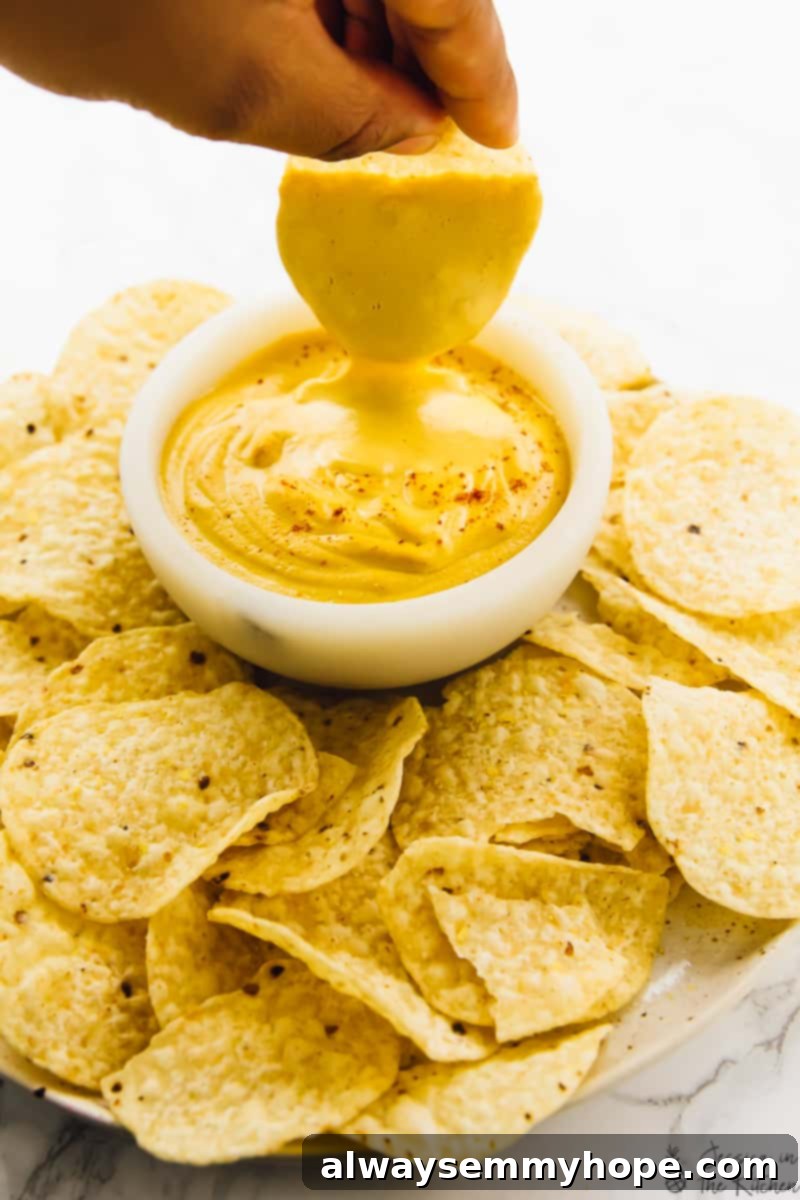 A hand enthusiastically dips a crispy tortilla chip into a small, inviting bowl of creamy vegan cheese sauce, surrounded by an abundance of other tortilla chips, setting the scene for a perfect snack or appetizer.