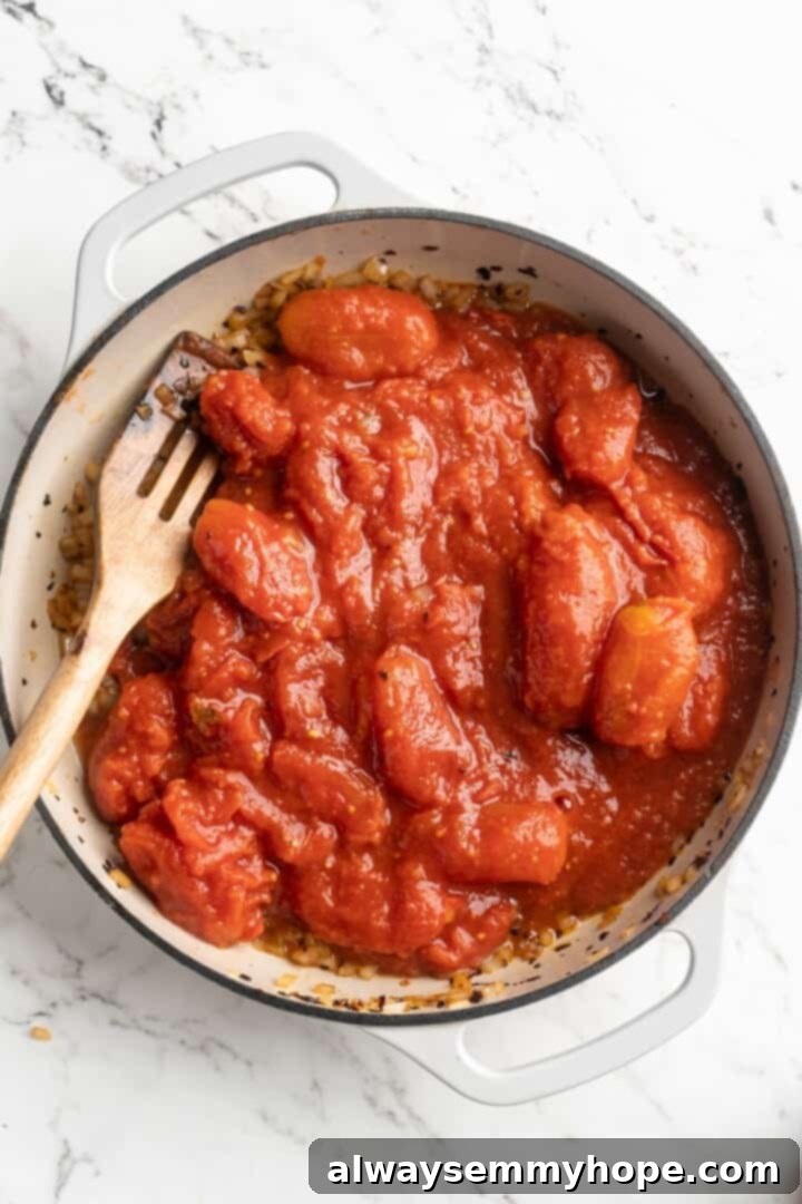 Overhead view of a slotted wooden spoon breaking down whole peeled tomatoes directly in the skillet during the initial simmer of arrabbiata sauce.