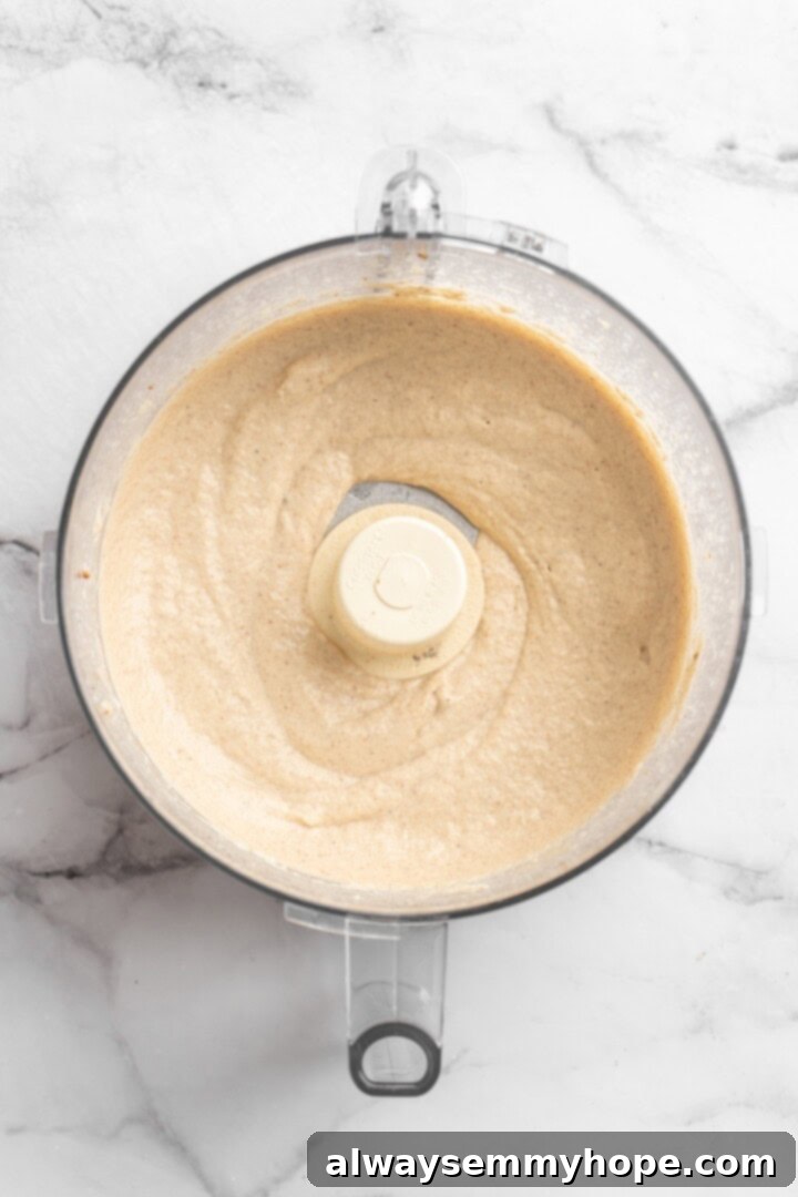 This easy Roasted Cauliflower Hummus gives the classic dip a low-carb makeover! I add roasted garlic too, which makes it extra delicious. Overhead view of roasted cauliflower hummus achieving a smooth and fluffy texture inside the food processor as tahini and ice-cold water are slowly incorporated