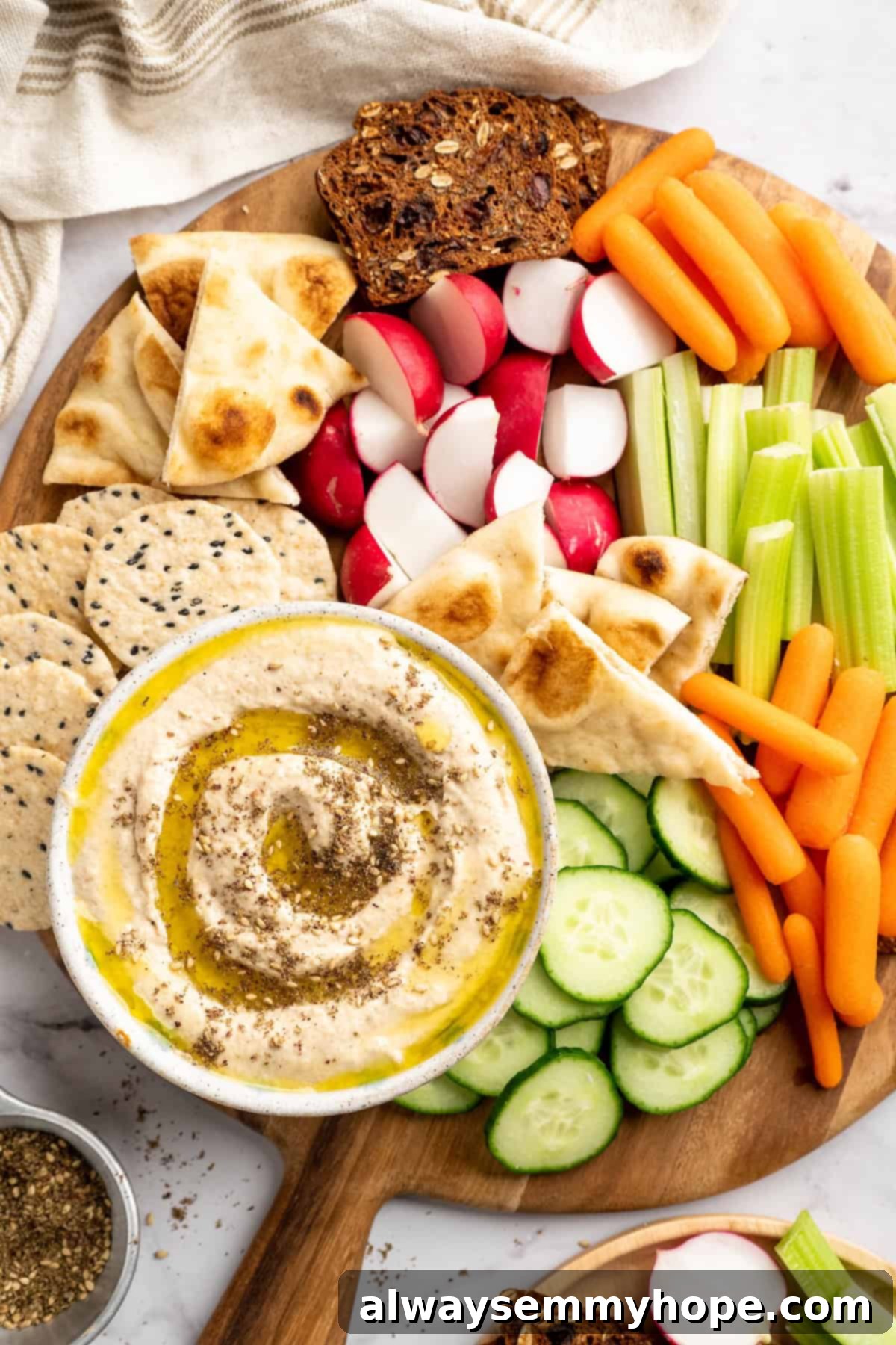 This easy Roasted Cauliflower Hummus gives the classic dip a low-carb makeover! I add roasted garlic too, which makes it extra delicious. Overhead view of a beautiful spread featuring a bowl of roasted cauliflower hummus, accompanied by fresh artisan bread, an assortment of crunchy crackers, and a selection of colorful raw vegetables like carrots, cucumbers, and bell peppers for dipping