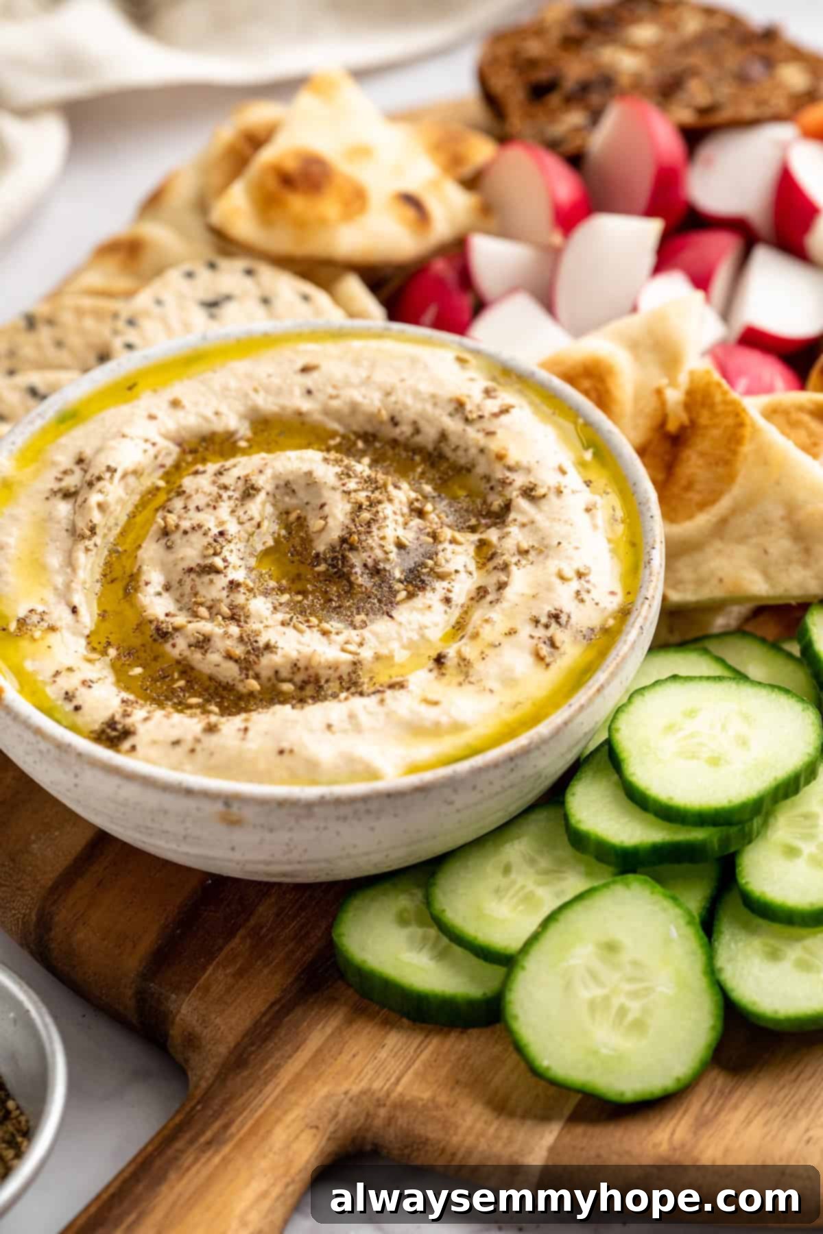 This easy Roasted Cauliflower Hummus gives the classic dip a low-carb makeover! I add roasted garlic too, which makes it extra delicious. A close-up shot of a bowl of roasted cauliflower hummus, garnished with fresh herbs and a drizzle of olive oil, displayed elegantly with a side of crisp, vibrant vegetables like cucumber slices and carrot sticks
