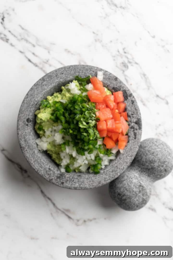 This easy guacamole recipe is the ultimate crowd pleaser! It’s incredibly fresh and flavourful, and it takes just 15 minutes to make. Overhead view of all guacamole ingredients, including mashed avocado, tomato, onion, and cilantro, mixing in a bowl