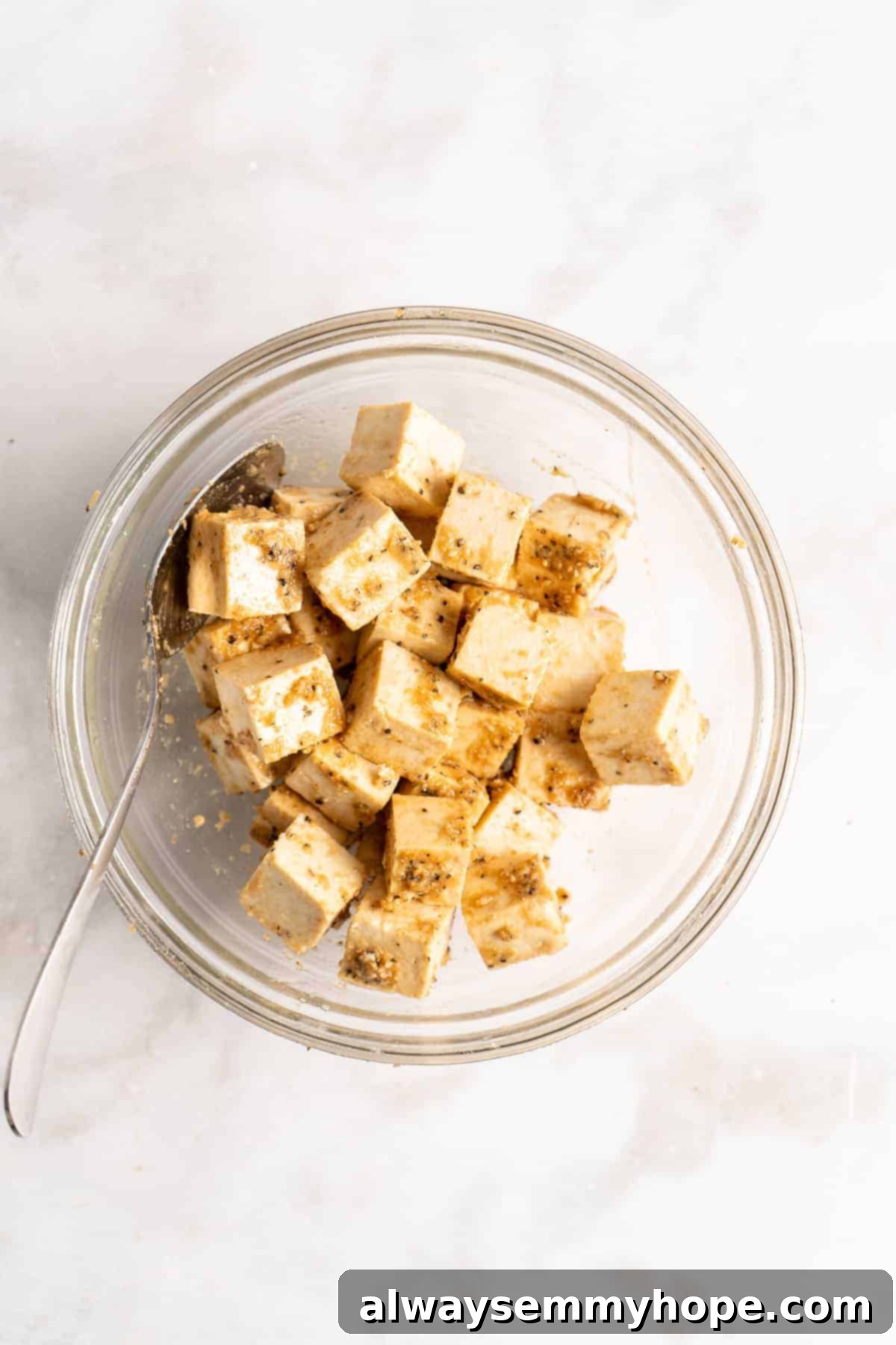 An overhead shot of pressed and cubed tofu in a glass mixing bowl, seasoned with liquid aminos and other spices, ready for air frying.