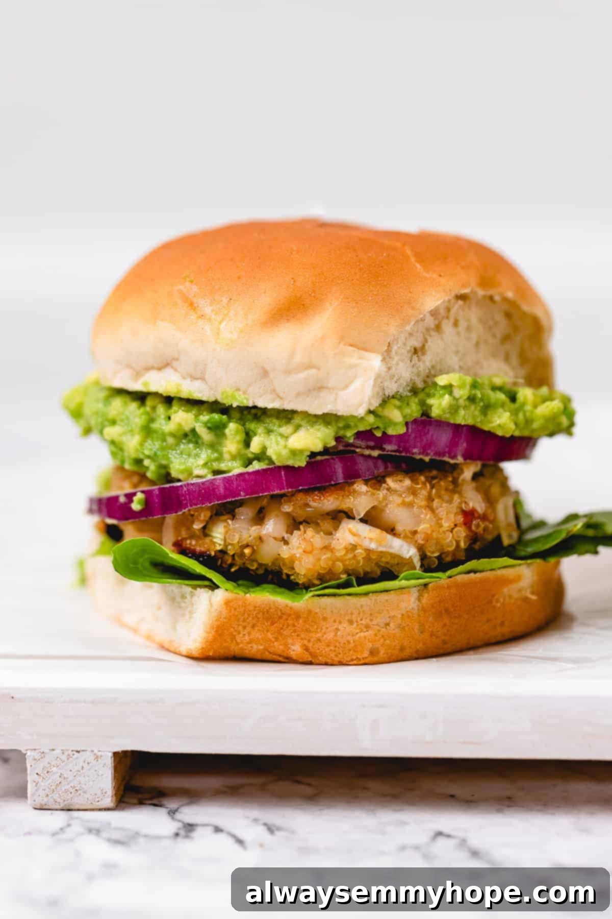 A close-up of a golden brown quinoa veggie burger with fresh lettuce, vibrant red onion, and creamy avocado slices on a white bun, served on a wooden board.