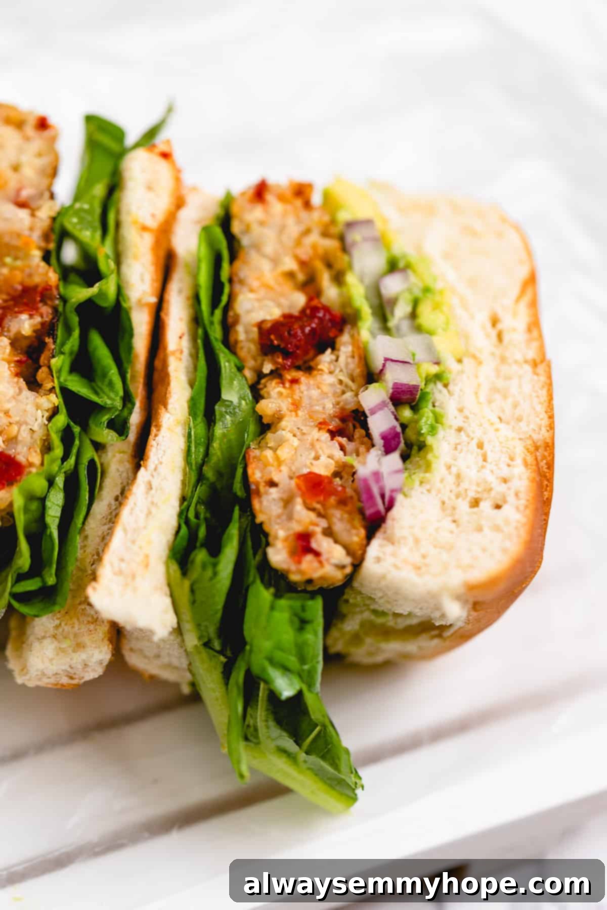 A cross-section view of a perfectly assembled vegan quinoa veggie burger, showing the layers of the golden patty, fresh red onion, crisp lettuce, and creamy avocado within a fluffy burger bun.