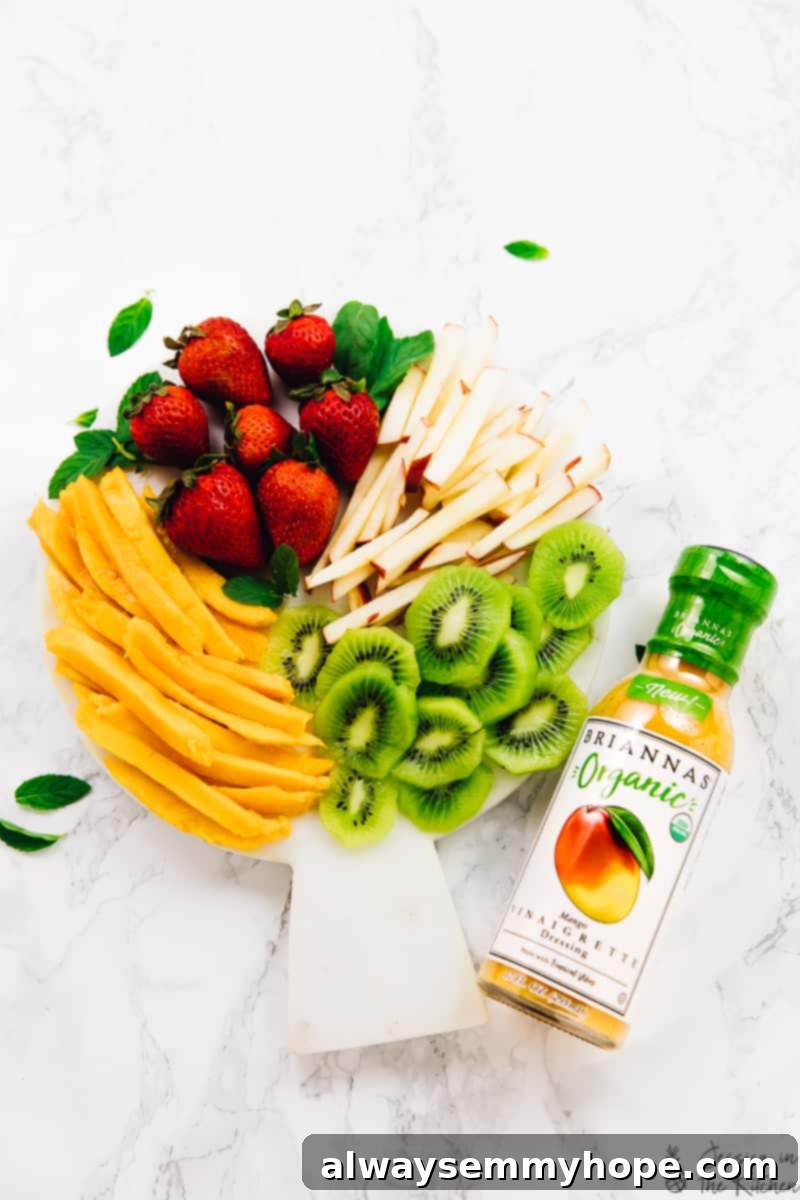 Tropical Fruit Spring Rolls with Sunshine Mango Dip 3 An overhead shot showcasing beautifully sliced fresh fruits, including mangoes, kiwis, strawberries, and apples, arranged artfully on a cutting board, ready to be wrapped into spring rolls. Next to them sits a bottle of creamy mango vinaigrette dressing, indicating the key ingredient for the accompanying dip. The scene highlights the fresh ingredients and the perfect pairing for the Summer Fruit Spring Rolls.