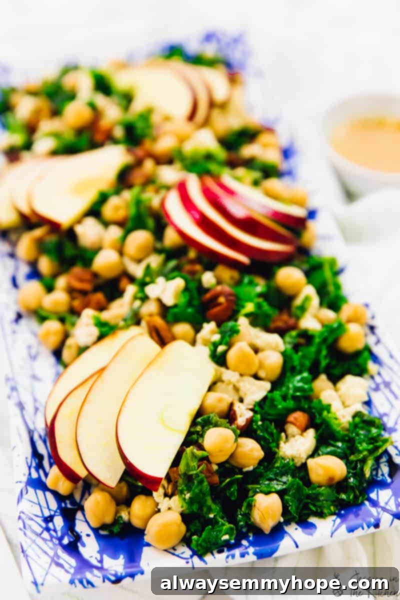 A close-up overhead shot of the finished kale apple salad, beautifully arranged on a rectangular platter, showcasing its vibrant colors and inviting textures with chickpeas, pecans, and vegan feta prominently displayed.