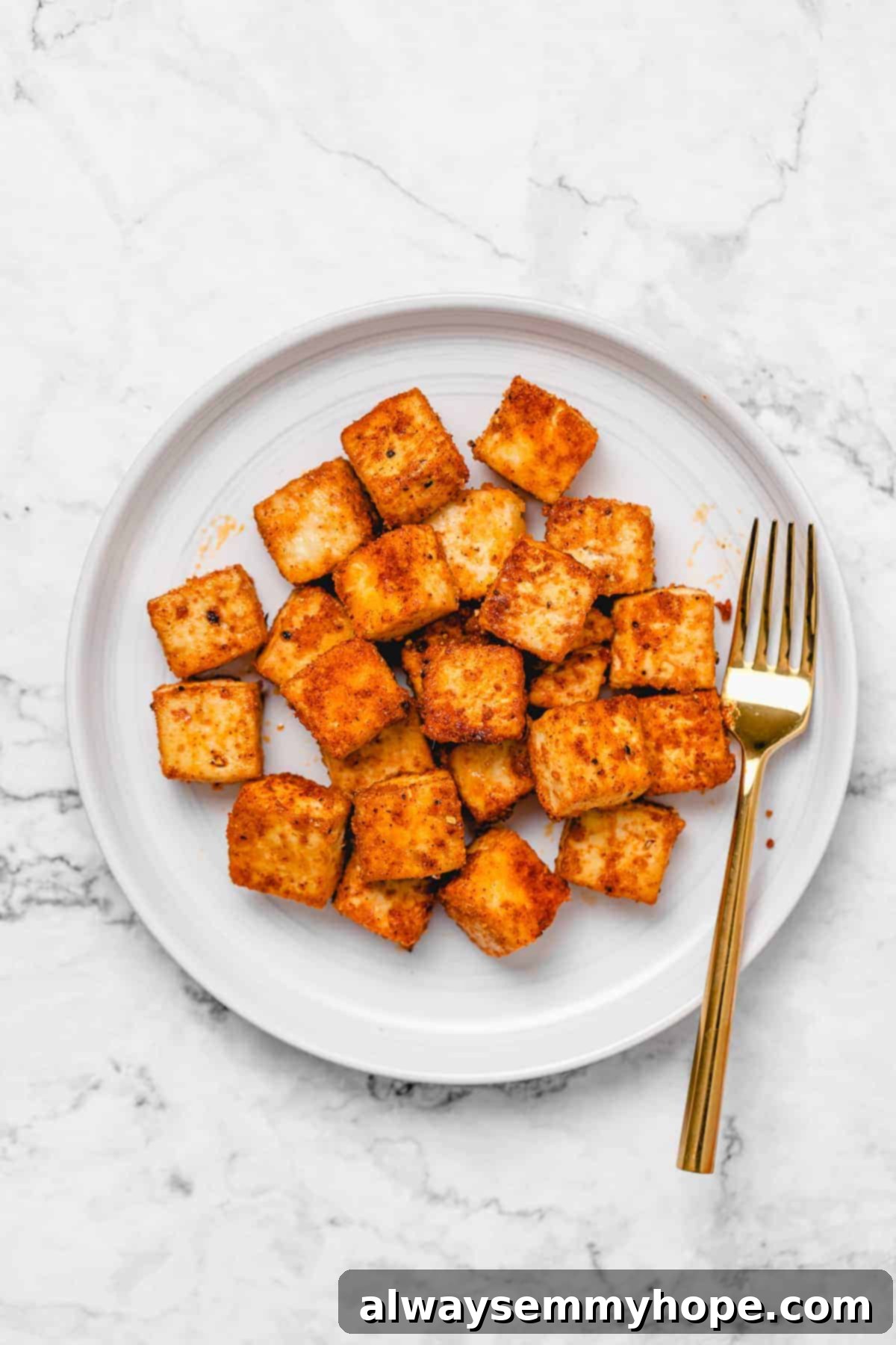 Achieve perfectly crispy and flavorful baked tofu with this simple 20-minute recipe, ideal for quick meals and meal prep! Crispy baked tofu cubes arranged artfully on a white round plate with a golden fork, highlighting their appealing texture.