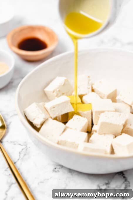 Tossing cubed tofu with oil and soy sauce for the initial marinade layer. Olive oil being poured over a bowl of cubed tofu, with light soy sauce already added.
