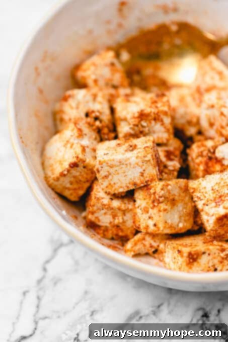 Adding and mixing dry seasonings to the tofu for a flavorful and crispy coating. Seasoned raw tofu marinating in a white bowl with a golden spoon, coated in cornstarch and spices.