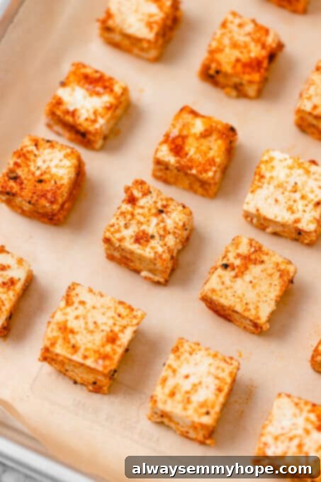 Arranging seasoned tofu in a single layer for optimal crisping in the oven. Cubes of uncooked firm tofu covered in seasoning, spread in a single layer on parchment paper on a baking sheet.