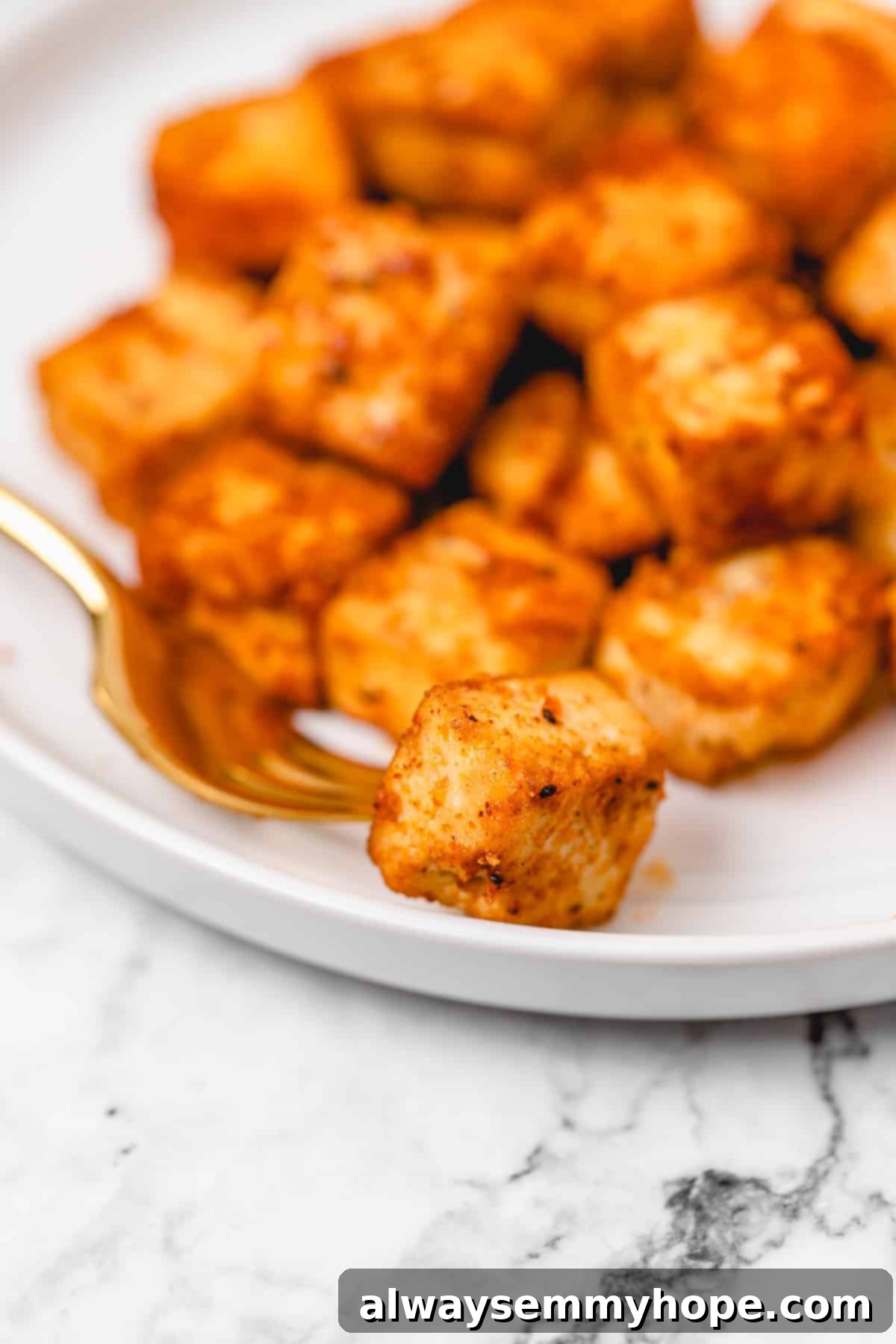 Enjoying a bite of delicious, perfectly crispy baked tofu. A perfectly crispy piece of baked tofu held on a gold fork, with a blurred plate of more baked tofu in the background.