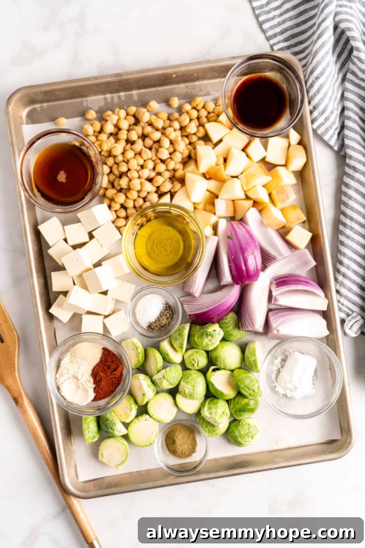 This vegan Buddha bowl is a flavor-packed meal brimming with caramelised roasted veggies, crispy chickpeas, tender baked tofu, and more! Overhead view of vegan Buddha bowl ingredients, including Brussels sprouts, red onion, and apple, arranged on a sheet pan before roasting.