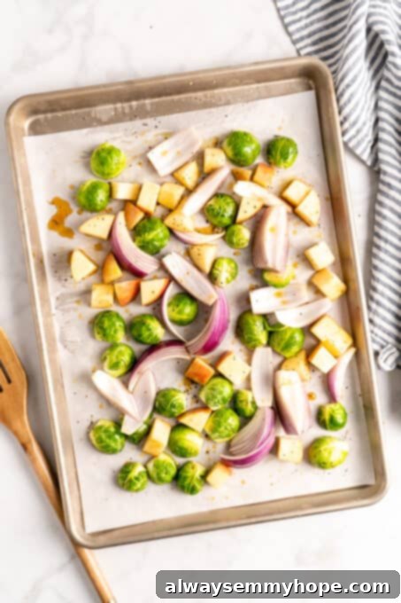 This vegan Buddha bowl is a flavor-packed meal brimming with caramelised roasted veggies, crispy chickpeas, tender baked tofu, and more! Overhead view of apples, Brussels sprouts, and red onions on a parchment lined sheet pan, ready for roasting.