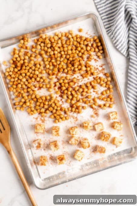 This vegan Buddha bowl is a flavor-packed meal brimming with caramelised roasted veggies, crispy chickpeas, tender baked tofu, and more! Overhead view of roasted chickpeas and tofu on a parchment lined sheet pan, golden brown and crispy.