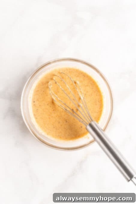This vegan Buddha bowl is a flavor-packed meal brimming with caramelised roasted veggies, crispy chickpeas, tender baked tofu, and more! Overhead view of spicy harissa tahini dressing in a glass bowl with a whisk.
