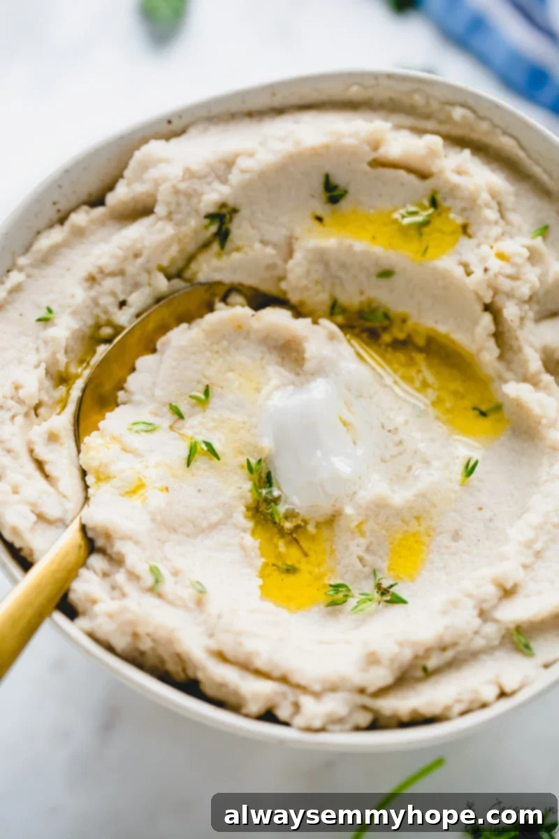 Closeup of mashed cauliflower in bowl with spoon, topped with melted vegan butter and fresh thyme leaves