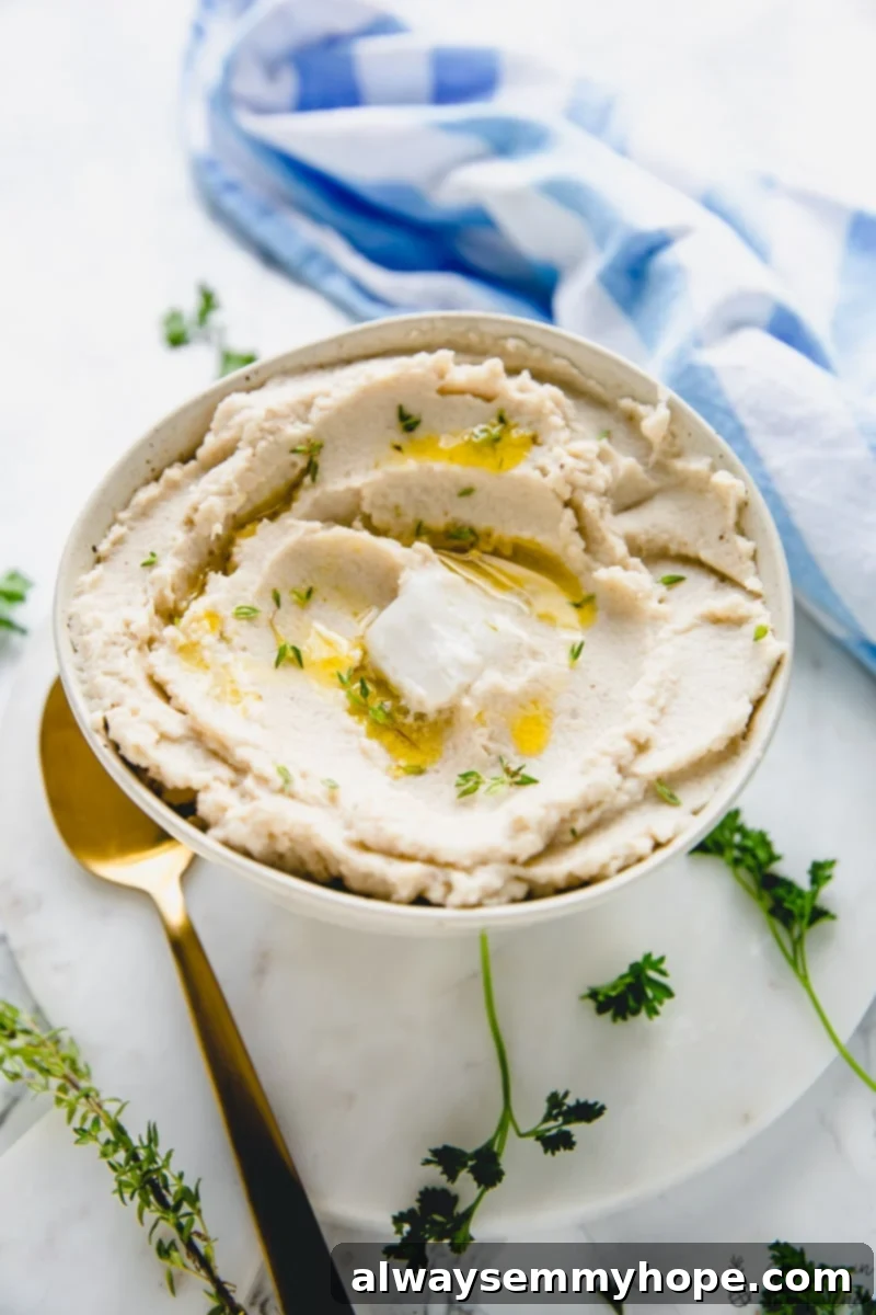 A bowl of cauliflower mash next to a blue cloth, with a gold spoon and sprigs of fresh herbs, ready to be served