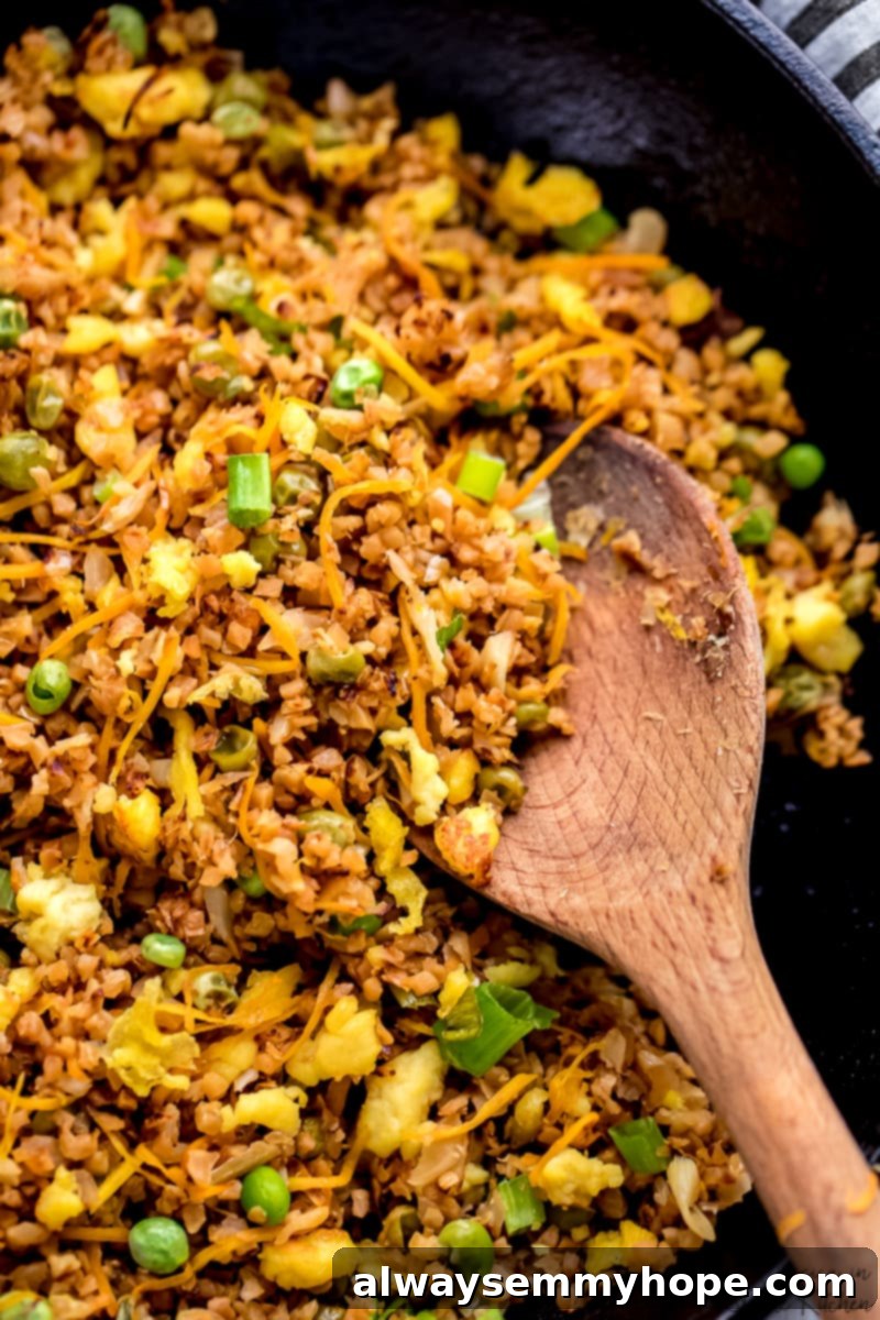A wooden spoon gently stirs the flavorful vegan fried rice mixture in a large black skillet, showcasing vibrant vegetables and cauliflower.
